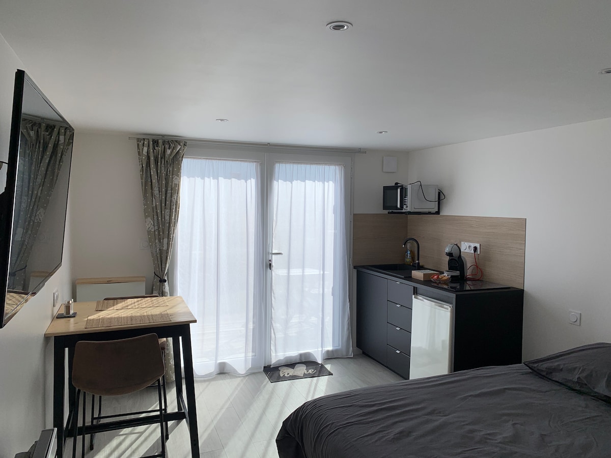 A studio apartment features a standard bed and a kitchenette with modern amenities, including a refrigerator and microwave. A small dining table is situated by large windows dressed with sheer curtains, allowing natural light to fill the space. A flat-screen TV is mounted on the wall.