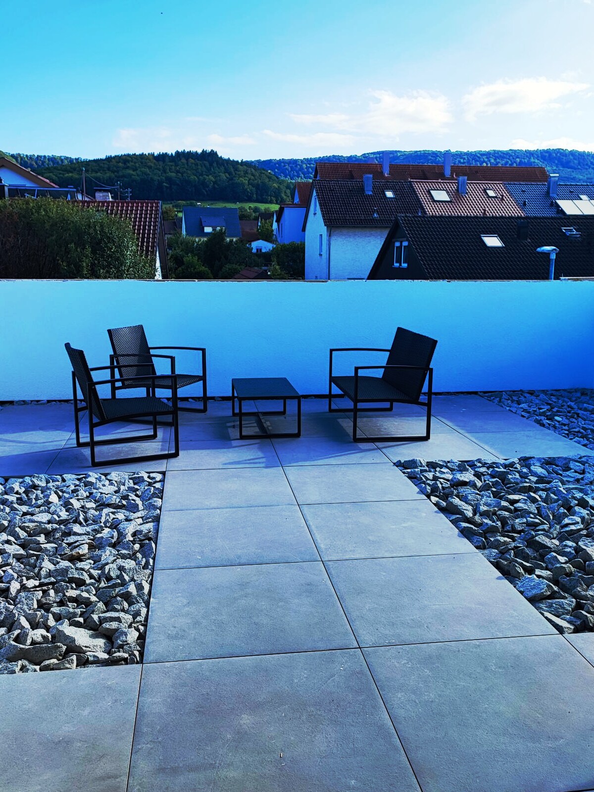 A small outdoor seating area is depicted, featuring four black chairs arranged around a rectangular table. The space is bordered by smooth stones and a white wall, with a view of nearby houses and rolling hills in the background under a clear blue sky.