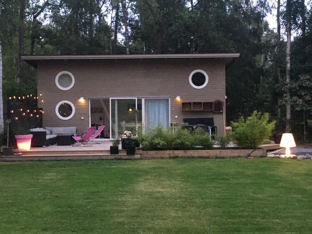 A modern house is set amidst a grassy area, featuring large circular windows and simple lines. The deck is furnished with a sofa, lounge chairs, and decorative lighting. A well-tended garden area with potted plants adds to the inviting exterior.