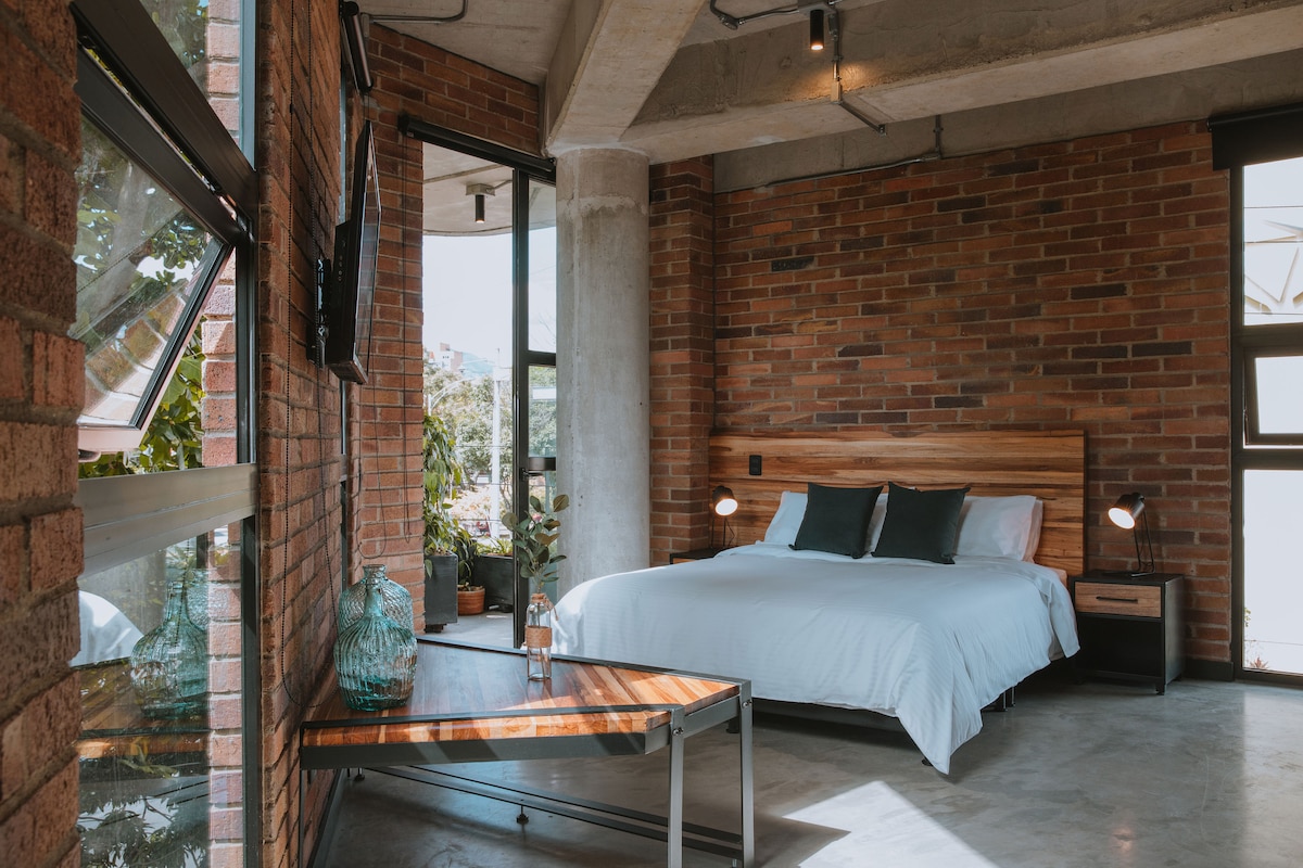 An open bedroom area features a spacious bed with crisp linens, complemented by two pillows. The exposed brick walls create a warm backdrop, while a modern metal table is positioned nearby. Large windows provide natural light and views of the surroundings.