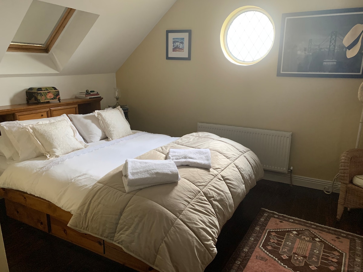 A cozy bedroom features a spacious bed adorned with soft white linens and neatly arranged towels. Natural light filters through an overhead skylight and a circular window. A wicker chair and decorative rug add warmth, while framed images enhance the room's character.