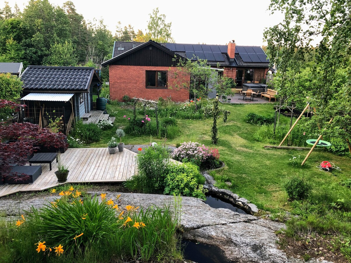 The garden features a variety of plants and flowers, including vibrant orange blooms. A wooden deck and stone path lead toward a spacious house with solar panels. A children's swing set and picnic area are visible, surrounded by lush greenery and trees.