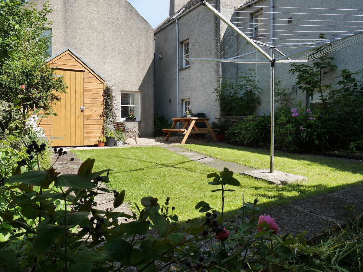 A secluded garden area features a well-maintained lawn bordered by plants and flowers. A wooden shed and a picnic table with benches are visible against the backdrop of the house. Sunlight bathes the space, enhancing its peaceful ambiance.