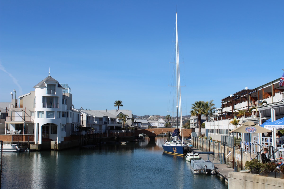 A serene canal view features a boat moored beside the walkway. Palm trees line the banks, and a bridge connects the waterfront buildings. The clear blue sky above complements the peaceful water, enhancing the relaxed waterfront atmosphere.