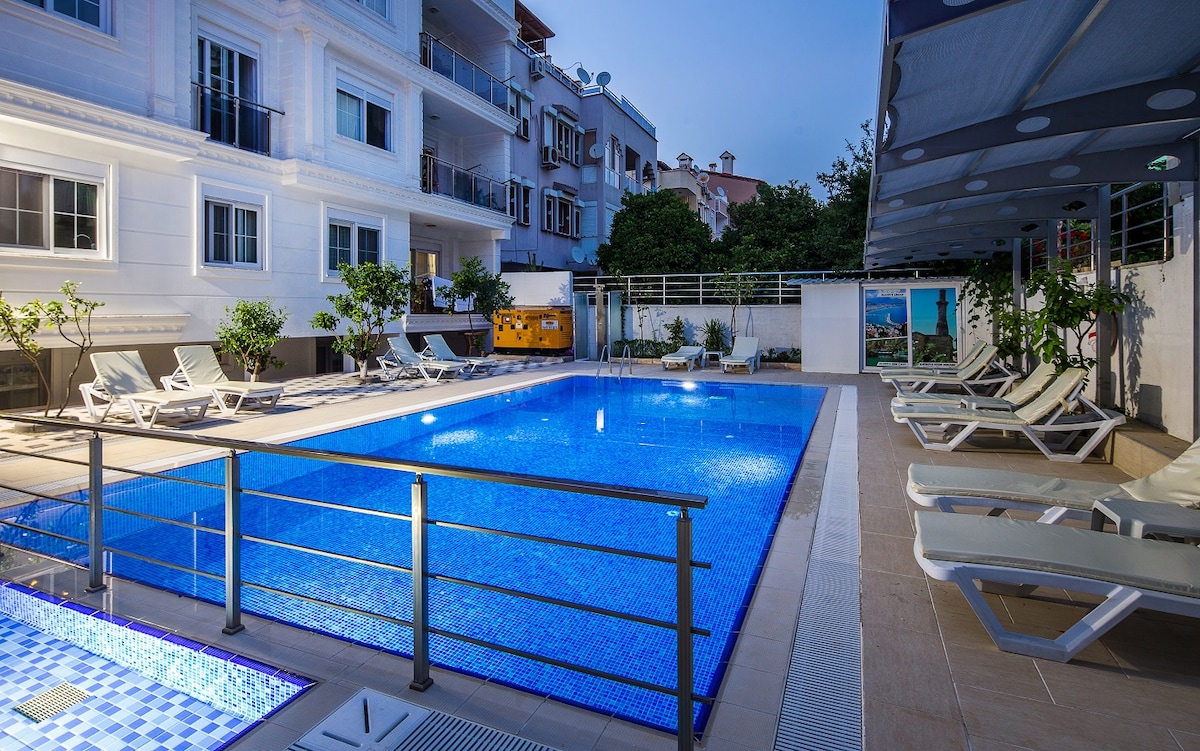 A swimming pool is set in a spacious outdoor area surrounded by lounge chairs. The pool features clear blue water with tile accents. Nearby, small trees are arranged along the perimeter, and a shaded seating area is visible in the background.
