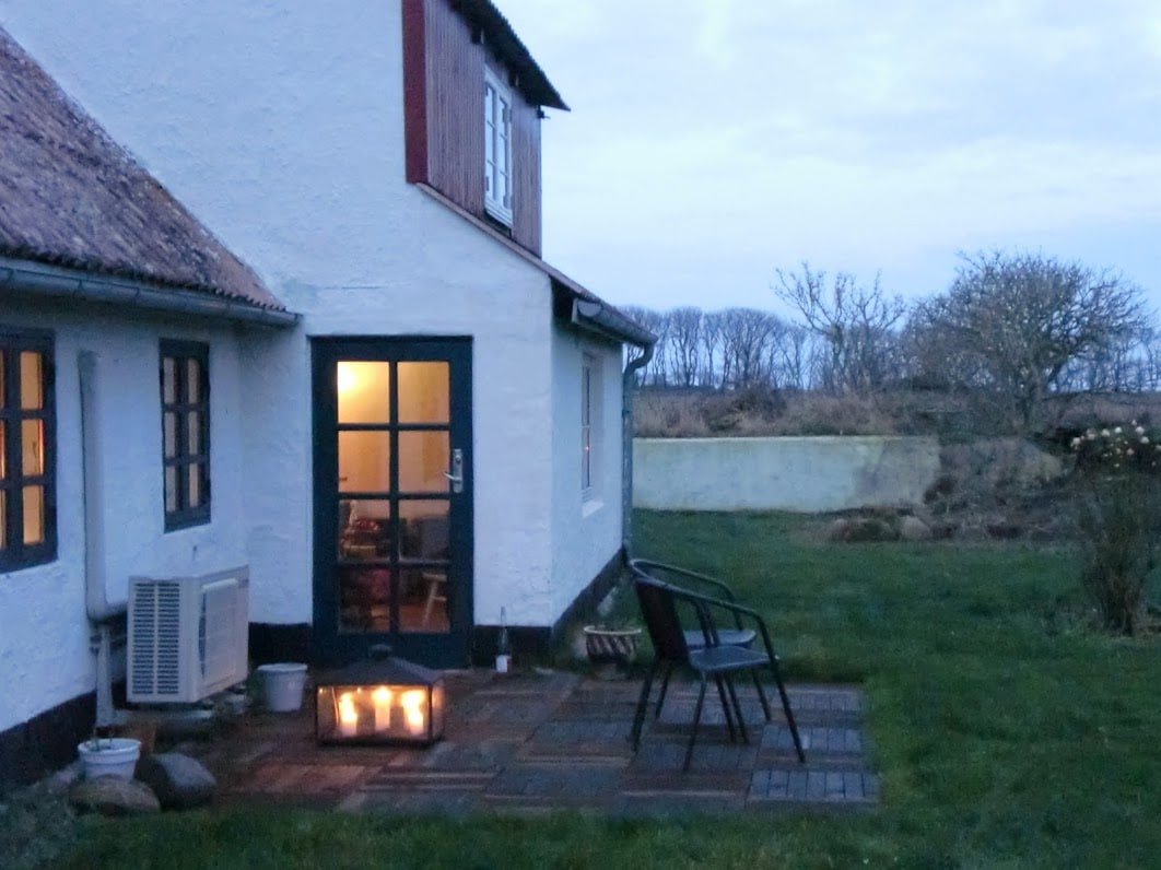 A quaint outdoor space is presented, featuring a small seating area with a chair and a lantern providing soft illumination. The house's white exterior and dark door contrast with the green grass surrounding the area. A glimpse of nature is visible in the background.