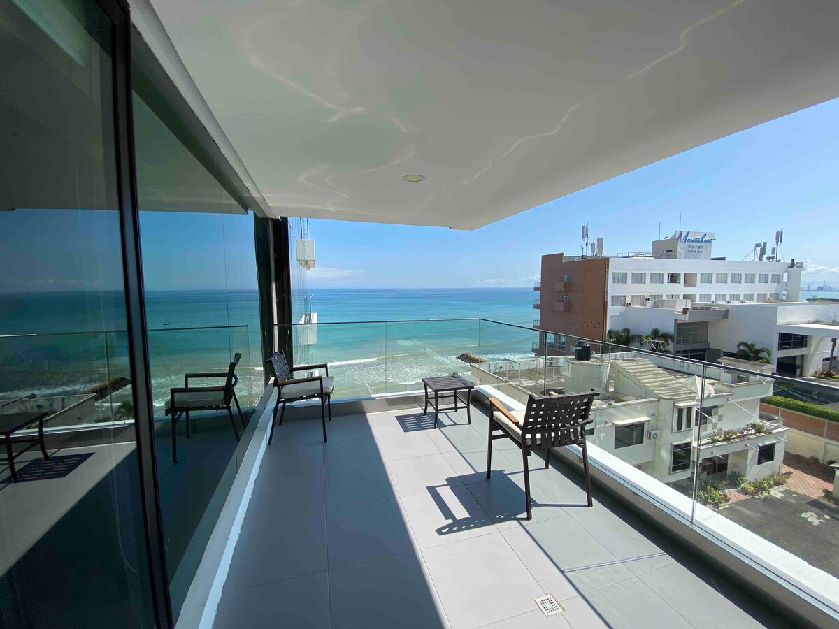 A spacious balcony offers panoramic views of the ocean and coastline. Elegant glass railings provide an unobstructed sightline, while two chairs and a lounge chair are positioned for relaxation. The bright sky and gentle waves are reflected in the nearby glass walls.