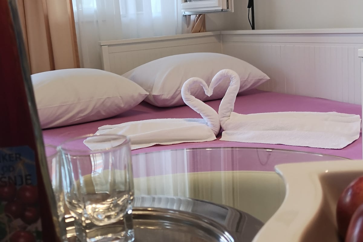 A neatly made bed features soft white pillows against a pastel purple cover. A towel arrangement shaped like swans is placed in the center, adding a touch of charm. In the foreground, a glass and a metal tray display fresh fruit, suggesting a welcoming atmosphere.