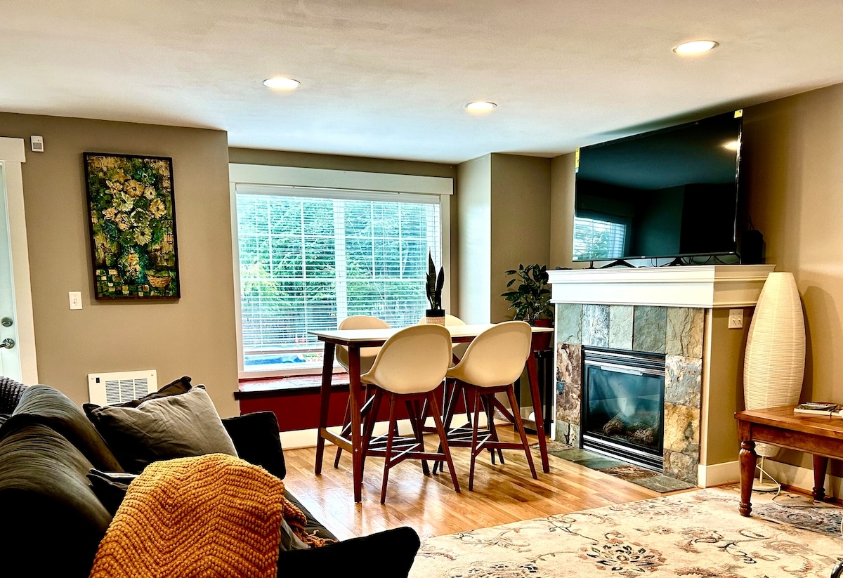 A cozy living area features a comfortable sofa and a round dining table set for four. Natural light fills the room through a large window. A fireplace adds warmth, while a mounted TV provides entertainment options. Decorated with a piece of wall art, the space is inviting.