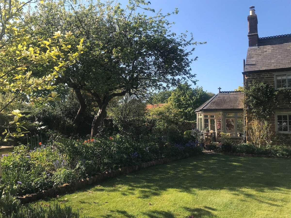 An inviting garden with well-maintained grass features a variety of colorful flowers and shrubs along the borders. A mature tree provides shade, while a cozy cottage is visible in the background, complementing the peaceful outdoor setting under a clear blue sky.