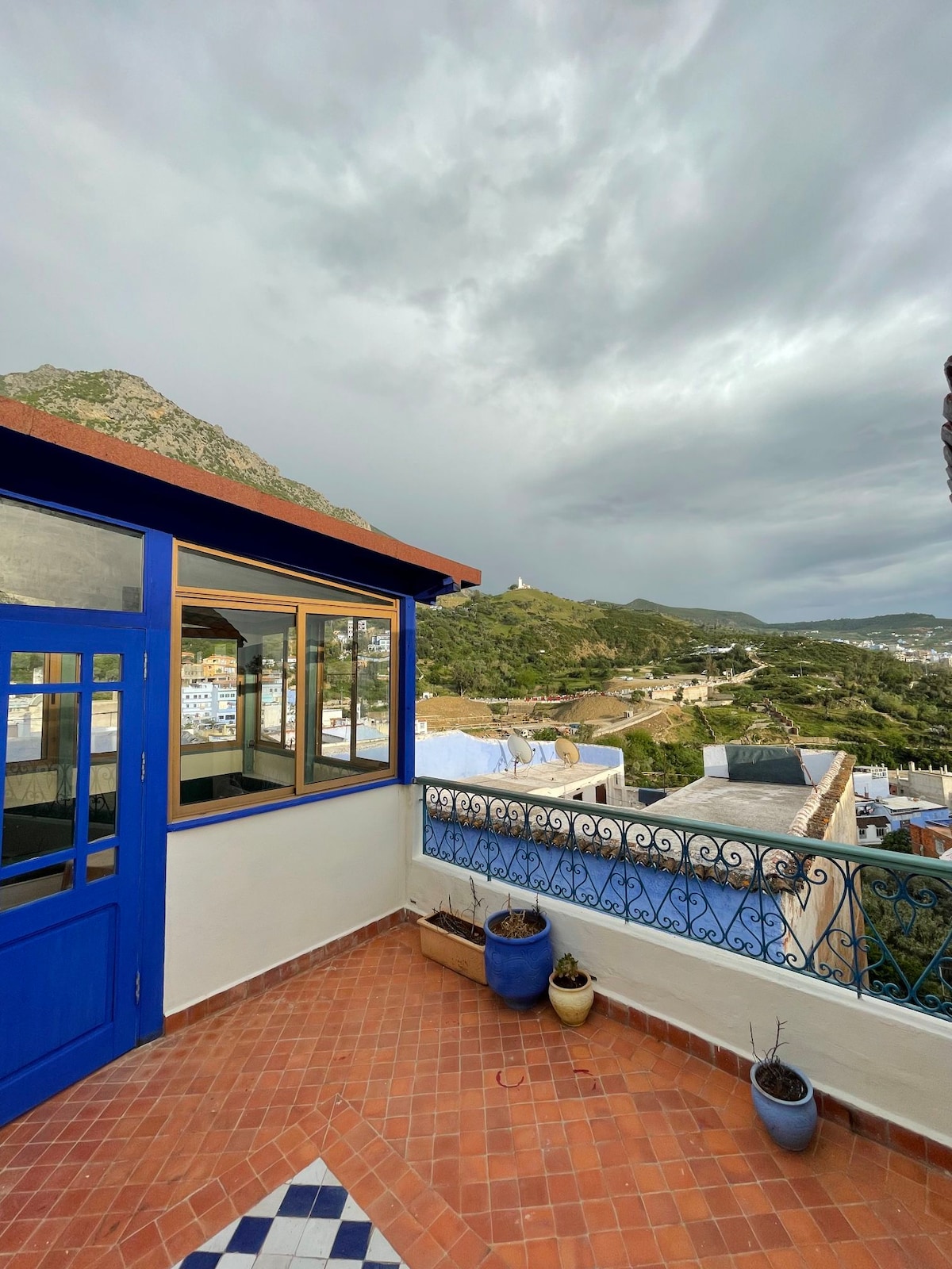 A spacious terrace is showcased with a striking blue railing and terracotta tiles. Panoramic views of a mountainous landscape and the surrounding blue city are visible. Large windows frame the area, allowing natural light to illuminate the inviting space.