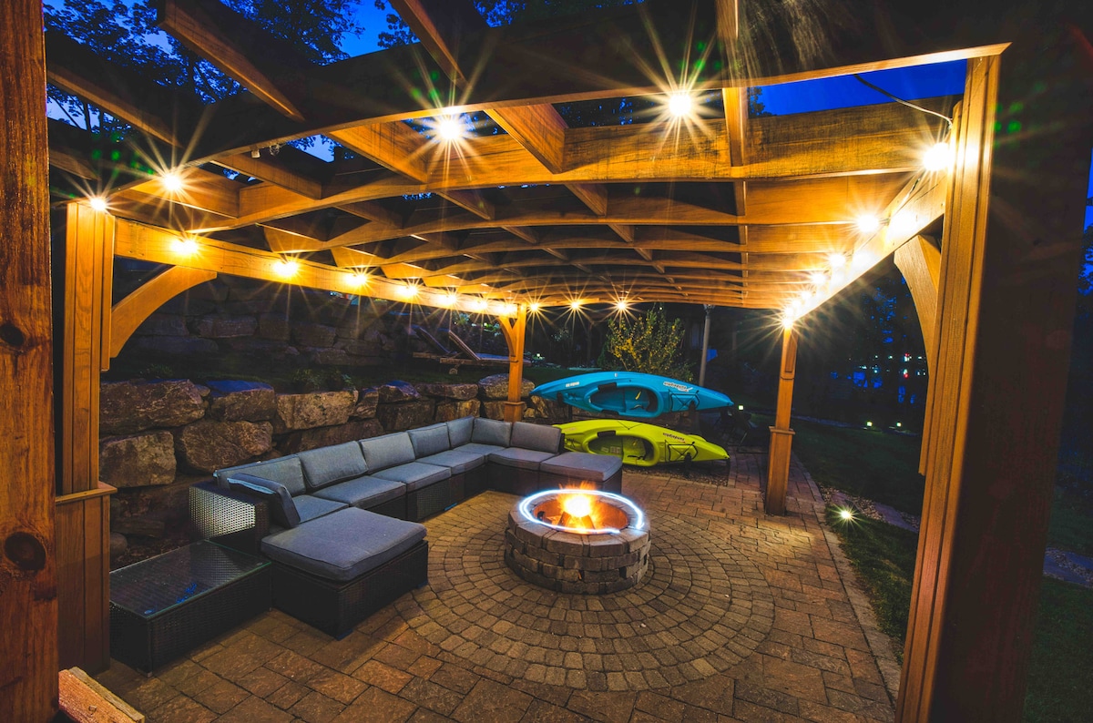 A spacious pergola is illuminated by string lights, creating a welcoming ambiance. A circular fire pit is surrounded by a stone seating area, with a large sectional couch offering ample seating. Two kayaks are stored nearby, hinting at outdoor adventures by the water.