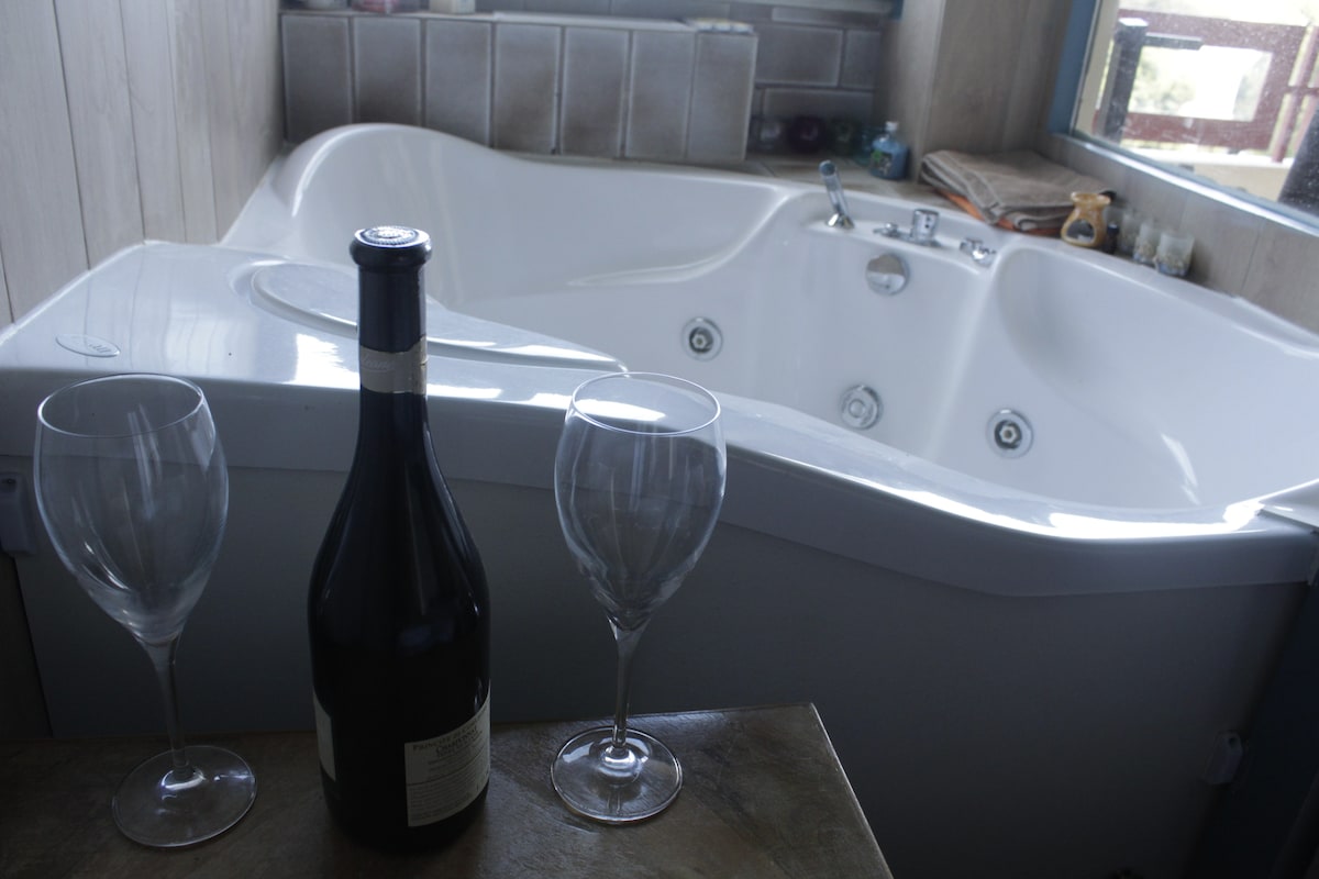 A spacious jacuzzi is featured in the image, accompanied by two clear wine glasses and a bottle of wine. Natural light filters through a nearby window, highlighting the elegant contours of the tub and the surrounding area.