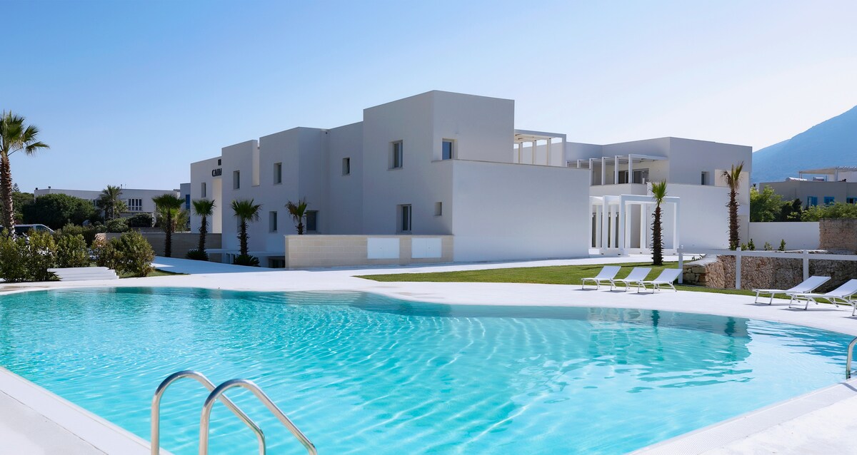 A modern resort complex is displayed, featuring a spacious swimming pool with clear turquoise water. Lush palm trees surround the pool area, and large white buildings are set against a bright blue sky, providing a tranquil and inviting atmosphere.