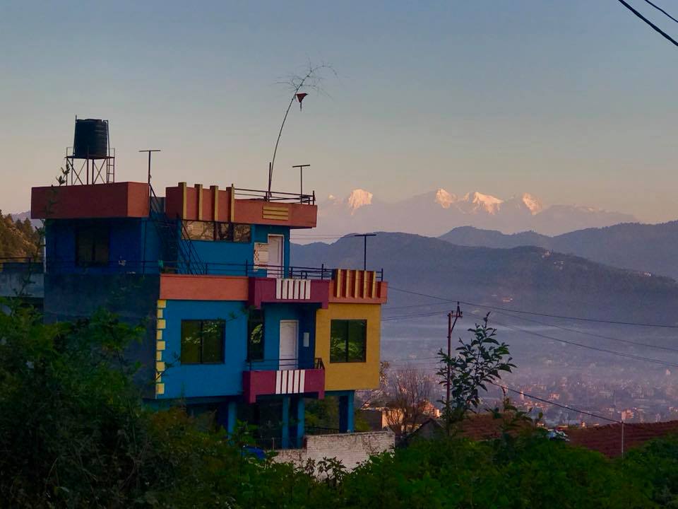 A colorful three-story house is set against a backdrop of distant snow-capped mountains. The building features vibrant hues and varied architectural elements, with windows and balconies visible on each level. Lush greenery surrounds the house, enhancing the natural setting.