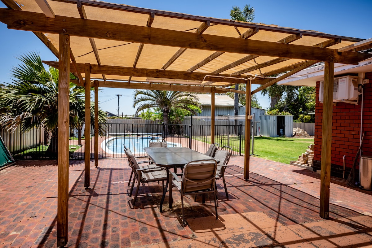 A shaded outdoor patio area features a large rectangular table surrounded by several chairs. The space is complemented by a swimming pool visible in the background, enclosed by a fence. Lush greenery and palm trees are present, enhancing the inviting setting.