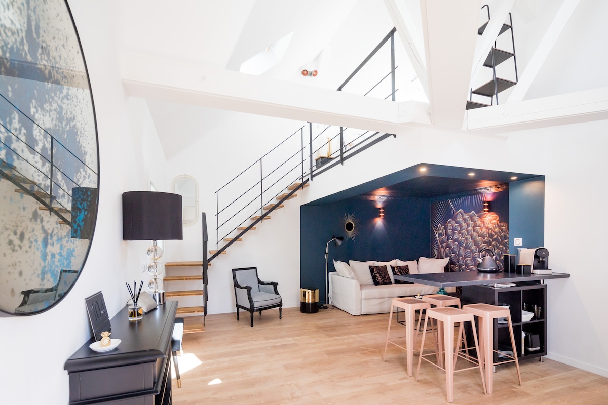 An open living space features high ceilings with exposed beams, promoting a sense of spaciousness. A staircase leads to an upper level. A modern seating area includes a light-colored sofa, accompanied by an armchair and minimalistic tables. A large round mirror enhances the room's depth.