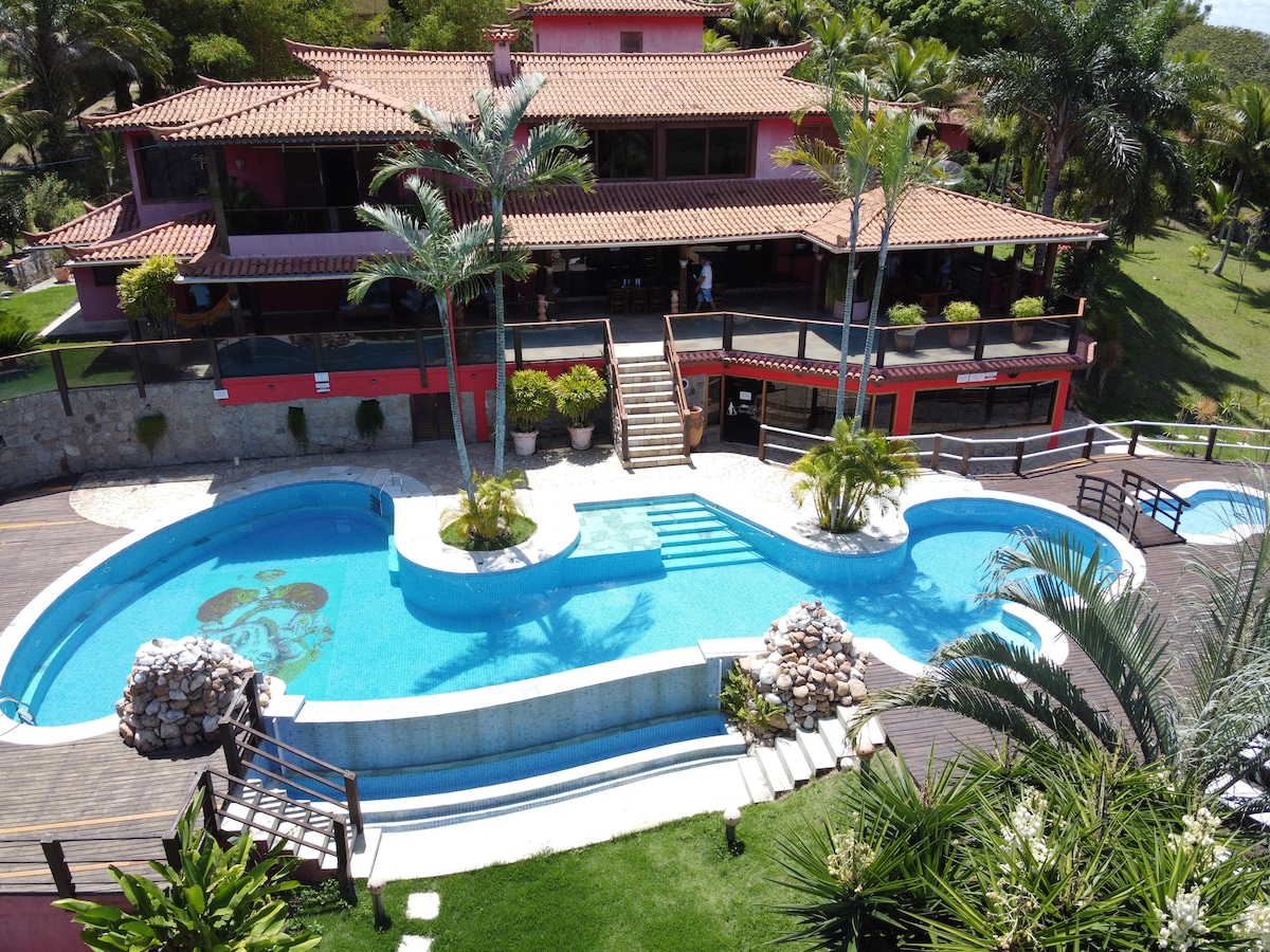 A spacious exterior view of the villa is presented, showcasing a large curved swimming pool surrounded by palm trees and tropical landscaping. The two-story structure features an expansive terrace with wooden railings, inviting connections to the lush green surroundings.