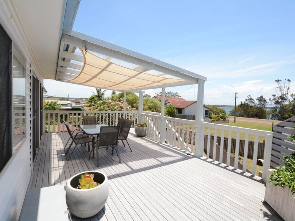 A spacious deck is featured, equipped with a dining table and chairs beneath a canopy for shade. The area offers expansive views of the surrounding landscape, with greenery visible in the distance and elements of coastal living captured in the serene setting.