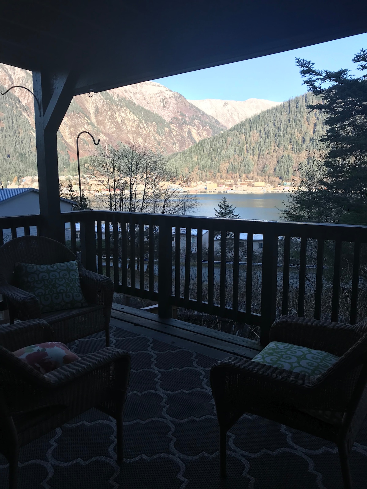A covered deck features two wicker chairs adorned with decorative pillows, offering a serene view of the mountains and water below. Natural light fills the space, highlighting the intricate patterns of the outdoor rug beneath.