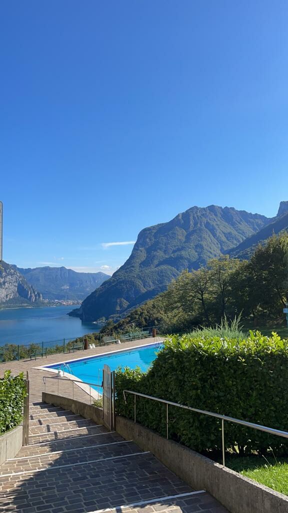 A view reveals a spacious rooftop pool surrounded by natural beauty, with lush greenery framing the area. The mountains rise majestically in the background, overlooking serene waters under a clear blue sky.