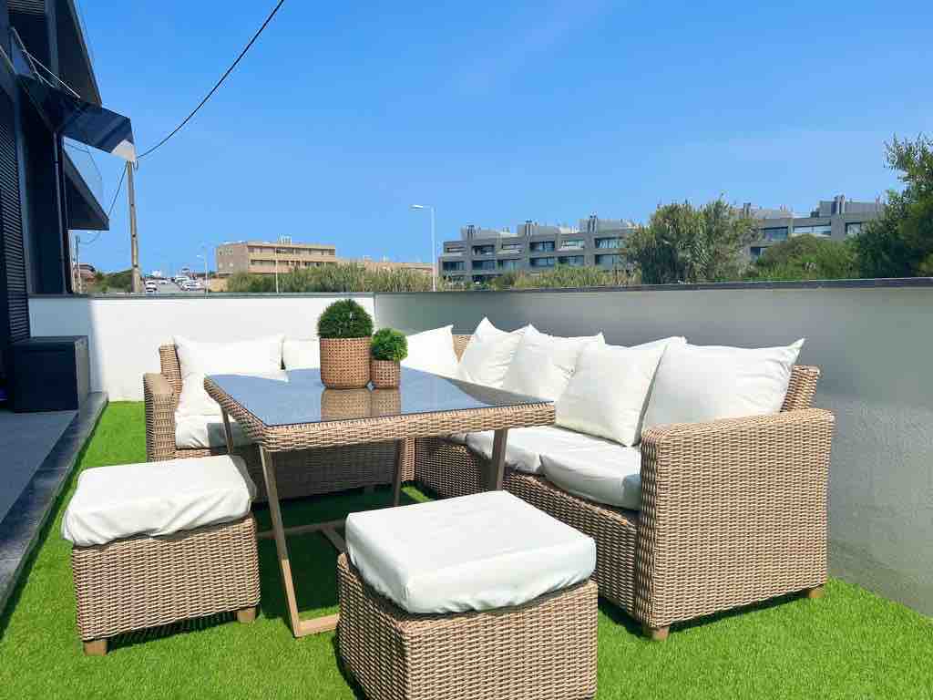 An outdoor seating area is furnished with a large wicker table and matching cushioned chairs. A few small ottomans are positioned nearby. The space is bordered by greenery and features an open, clear sky above.