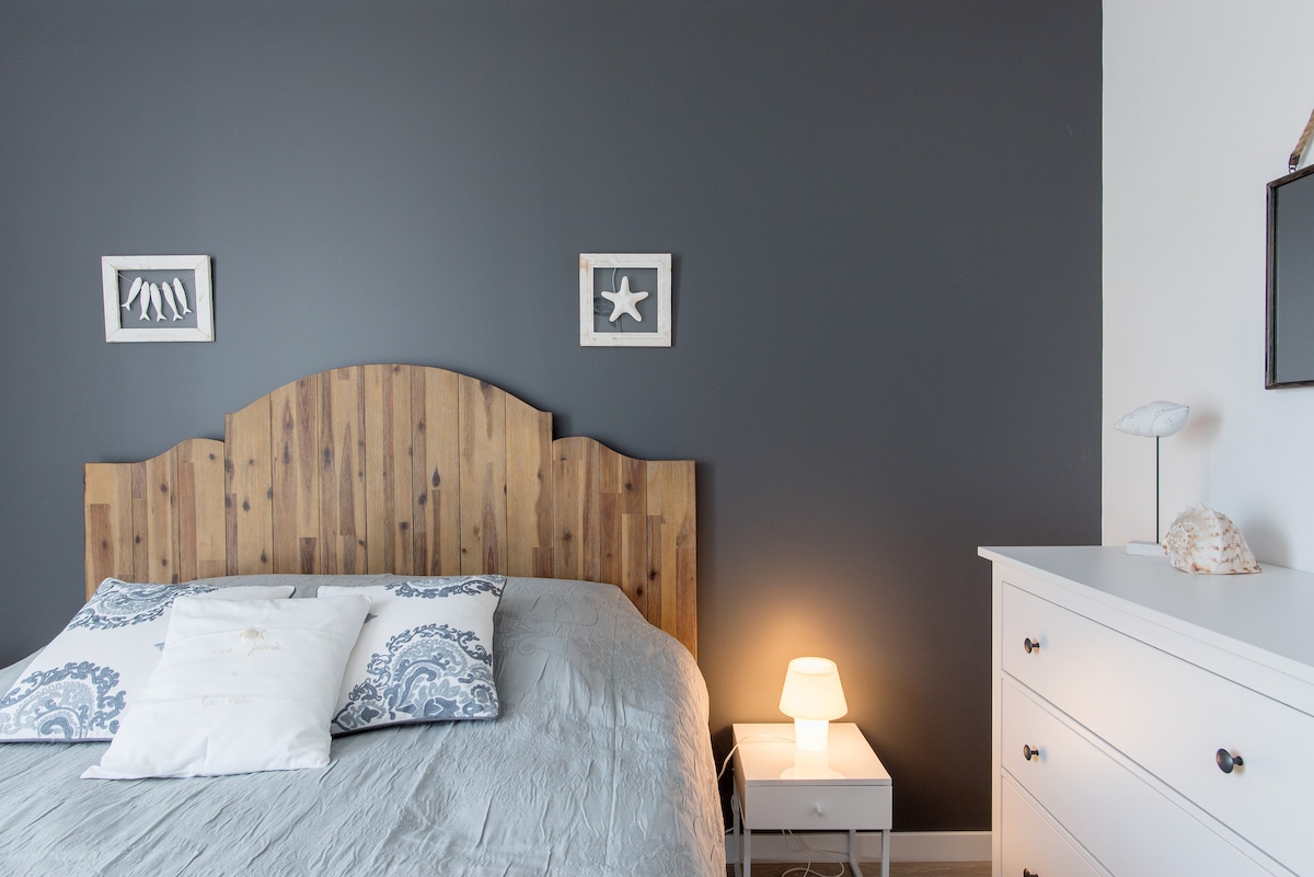 A cozy bedroom showcases a wooden headboard above a spacious bed dressed in soft linens. Light illuminates a small nightstand beside the bed, where a lamp and decorative elements are placed. A neutral wall provides a calming backdrop to the room.