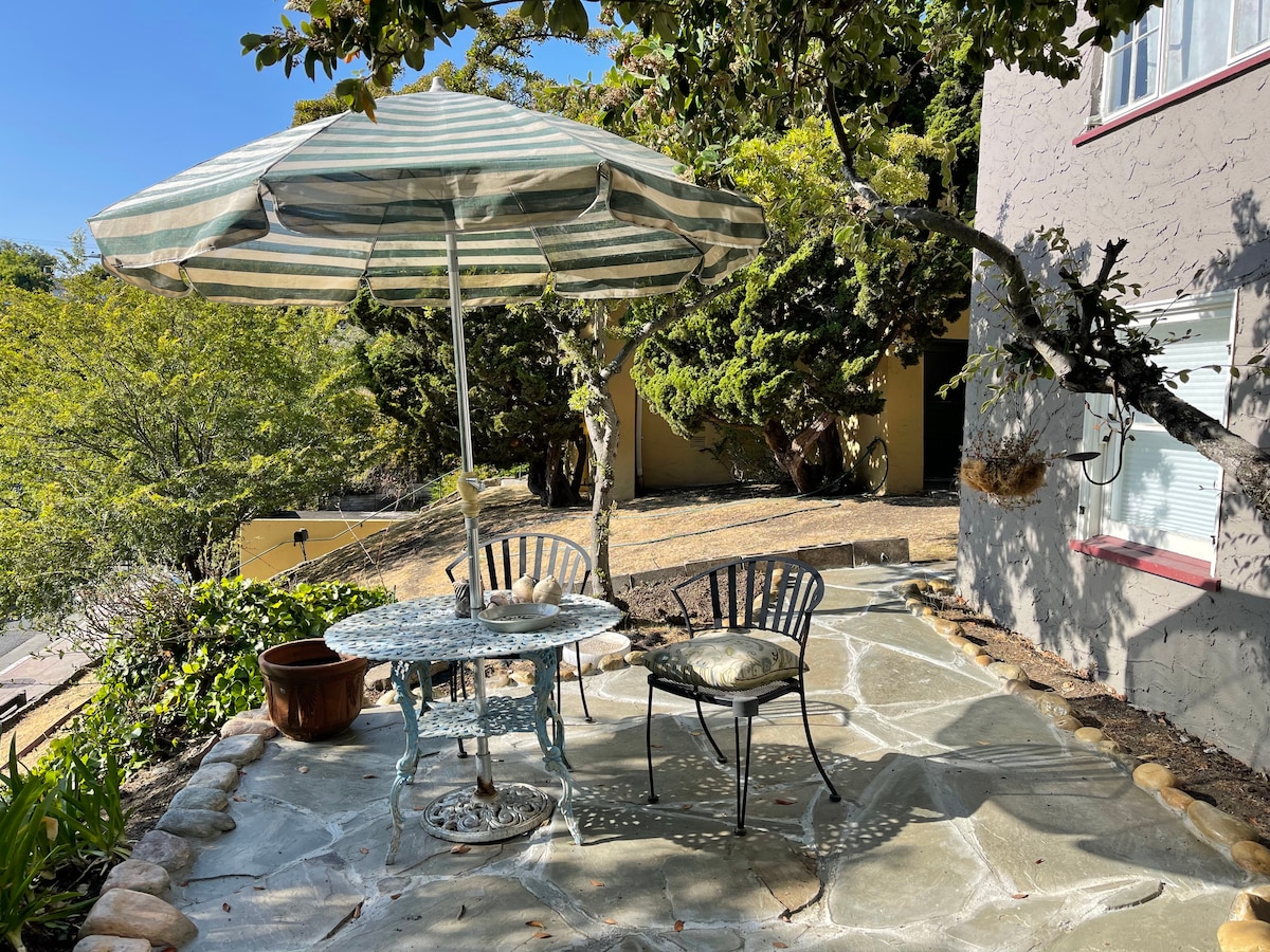 A shaded outdoor patio features a round table with a striped umbrella and two metal chairs, one with a cushion. The stone floor adds texture to the space, surrounded by greenery and trees, providing a peaceful setting for dining or relaxation.