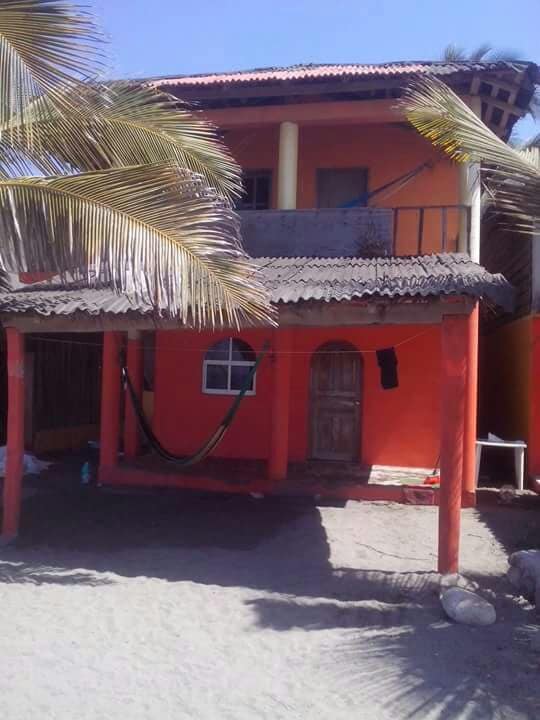 A vibrant two-story structure stands on sandy ground, featuring a bright orange façade and a thatched roof. A hammock is suspended between two posts, while a small window adds natural light to the entryway. Lush palm fronds frame the scene, enhancing the beachside setting.