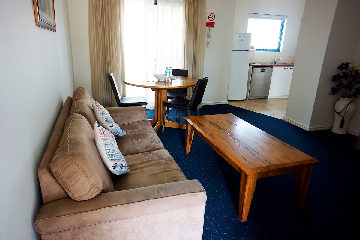 A comfortable living space features a plush sofa with decorative cushions, a wooden coffee table, and a round dining table set for four. Natural light filters through the sheer curtains, illuminating the open layout that includes a kitchenette visible in the background.