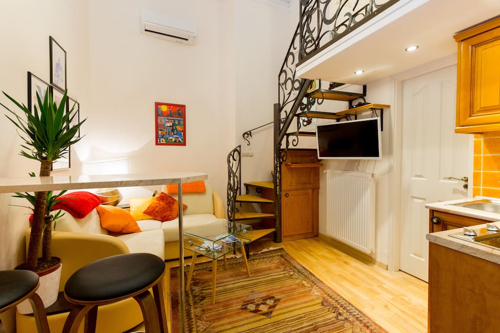 Dining area to the left, living room area with couch further down to the left, kitchen on the very right and white door entering onto the bathroom. Spiral staircase leading up to the mezzanine.