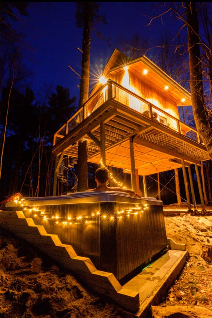 A treehouse in Maine illuminated from within at night, with a person enjoying the hot tub below