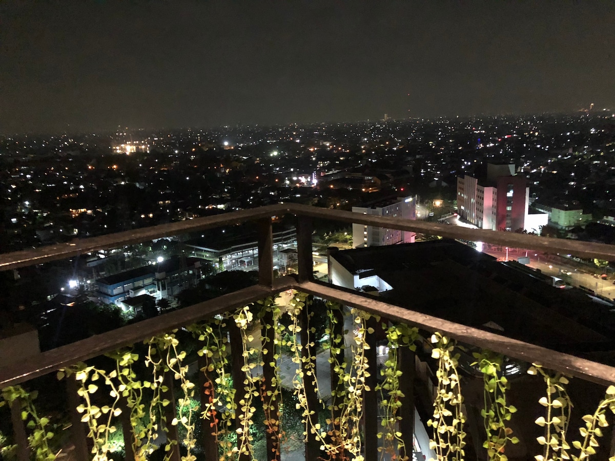 A balcony view at night showcases a sprawling city illuminated by countless lights. The railing is adorned with intertwining green vines, enhancing the serene backdrop of the urban landscape. Distant buildings and streets can be seen among the sparkling lights.