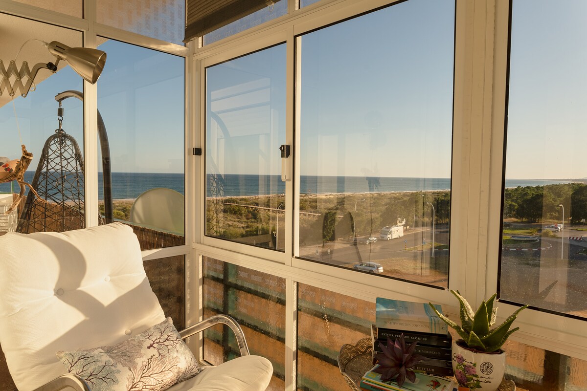 A bright sunroom features large glass windows offering unobstructed views of the ocean. A cozy white chair sits adjacent to the window, complemented by a decorative pillow. Potted plants add a touch of greenery to the space, creating an inviting atmosphere.