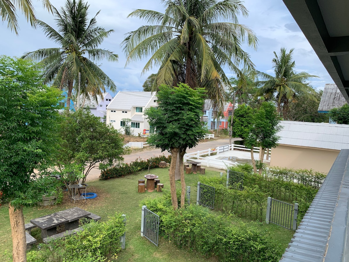 A view from a balcony showcases a green, well-maintained garden area featuring palm trees and bushes. Picnic tables are visible among the greenery. Colorful houses line the street in the background, creating a quaint and peaceful atmosphere.