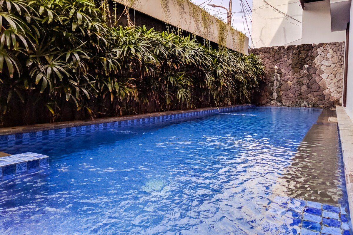A tranquil swimming pool is surrounded by lush greenery, with vegetation lining the walls. The clear blue water reflects the sky, creating a serene atmosphere. The pool area is designed for relaxation and enjoyment, offering a refreshing escape within the compound.