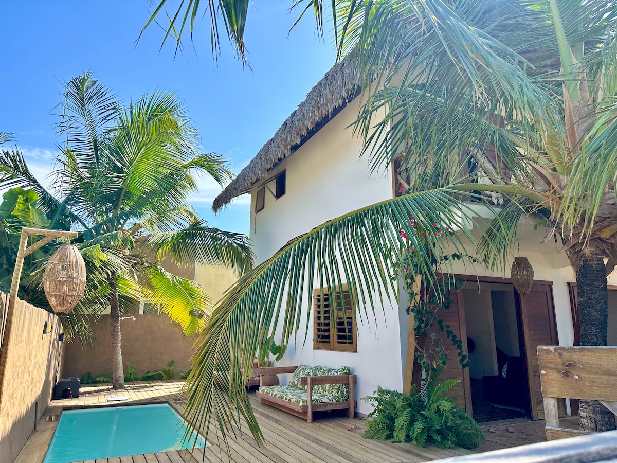 A stylish beach house is framed by lush palm trees, featuring a private swimming pool surrounded by wooden decking. The thatched roof adds a tropical touch, while outdoor seating invites relaxation in the sun. Light filters through the greenery, creating a vibrant atmosphere.