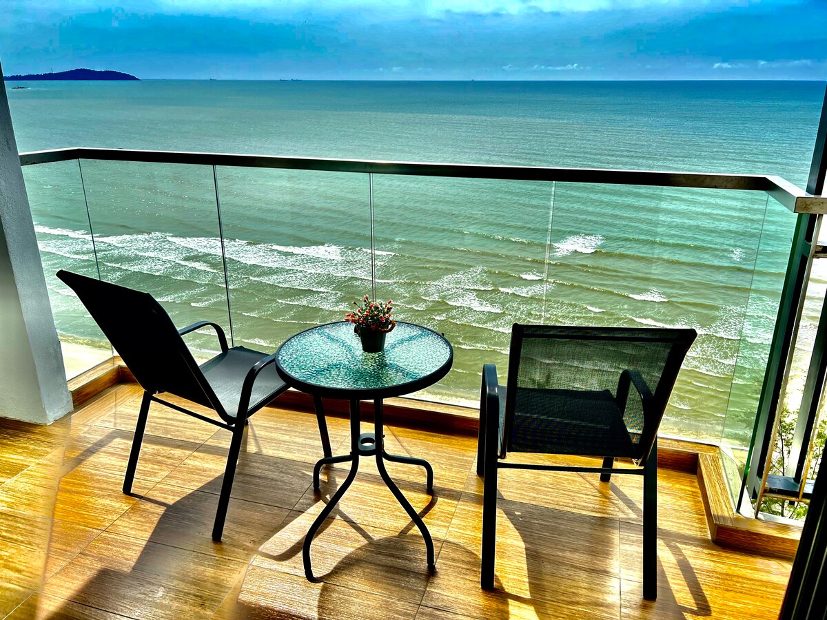 A balcony overlooks a scenic beachfront, featuring two lounge chairs positioned beside a small round table. A potted plant adds a touch of greenery, while the sea and waves can be seen in the distance under a clear sky.