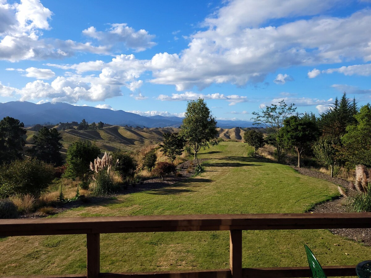 An extensive view of rolling hills stretches into the distance, framed by clear blue skies and fluffy white clouds. Various trees and shrubs punctuate the landscape, providing a natural setting that invites relaxation and contemplation.