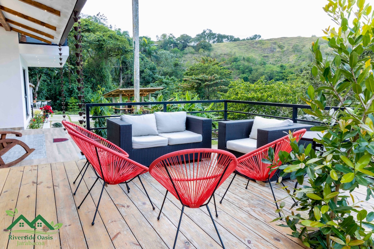 An outdoor seating area features comfortable cushioned sofas and four vibrant red chairs arranged around a wooden deck. Lush greenery and distant hills can be seen in the background, creating a serene environment for relaxation.