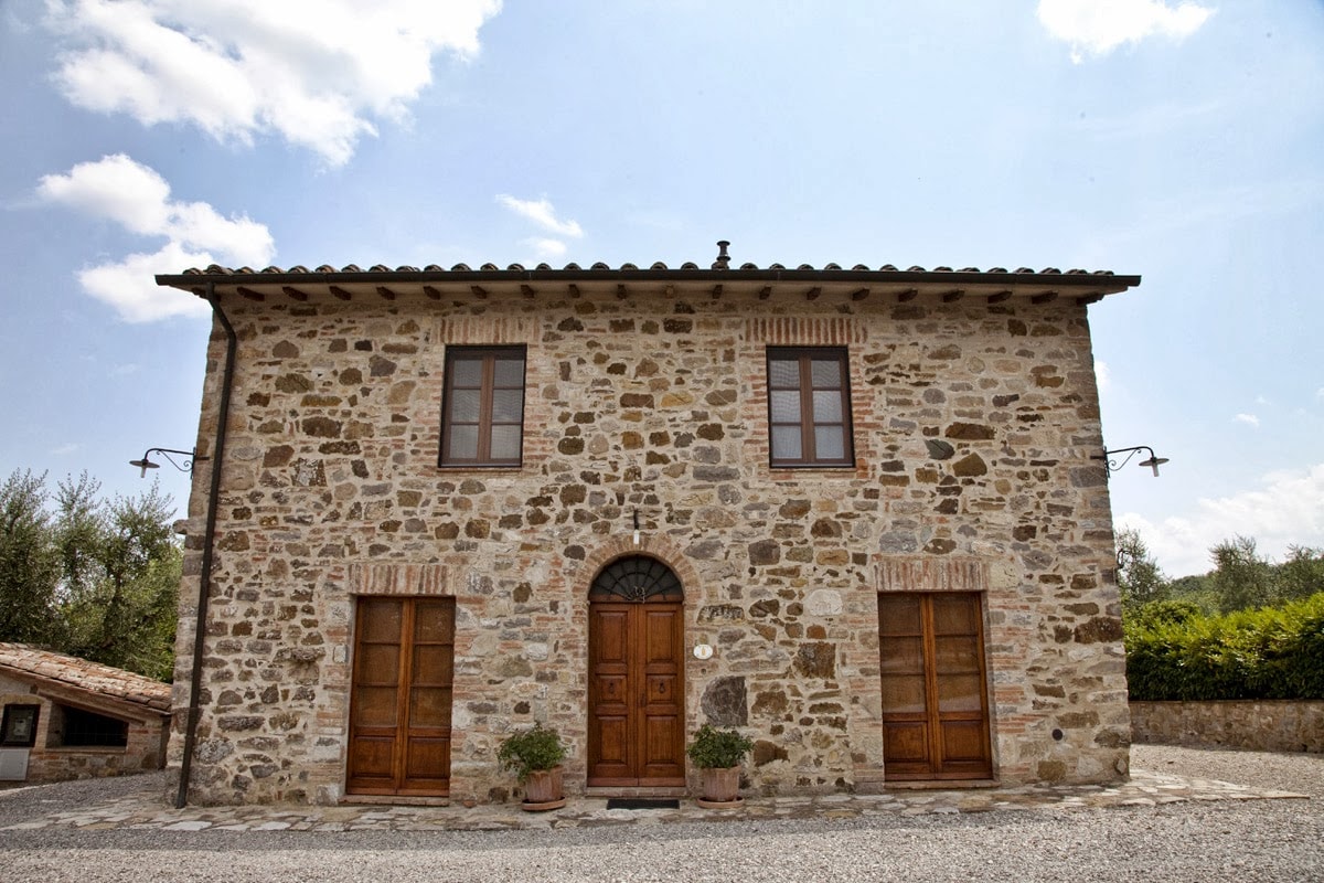 A traditional stone farmhouse stands prominently under a clear blue sky. The structure features large wooden doors framed by two sets of windows on the ground floor. Stone walls showcase natural textures. The surrounding gravel area is complemented by greenery in the background.