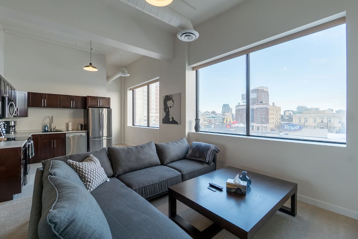 A spacious living area is highlighted by large windows that offer views of downtown Winnipeg. A comfortable sectional sofa is positioned beside a dark wooden coffee table, with an artistic wall piece visible nearby. The modern kitchen area features sleek cabinetry and stainless steel appliances.
