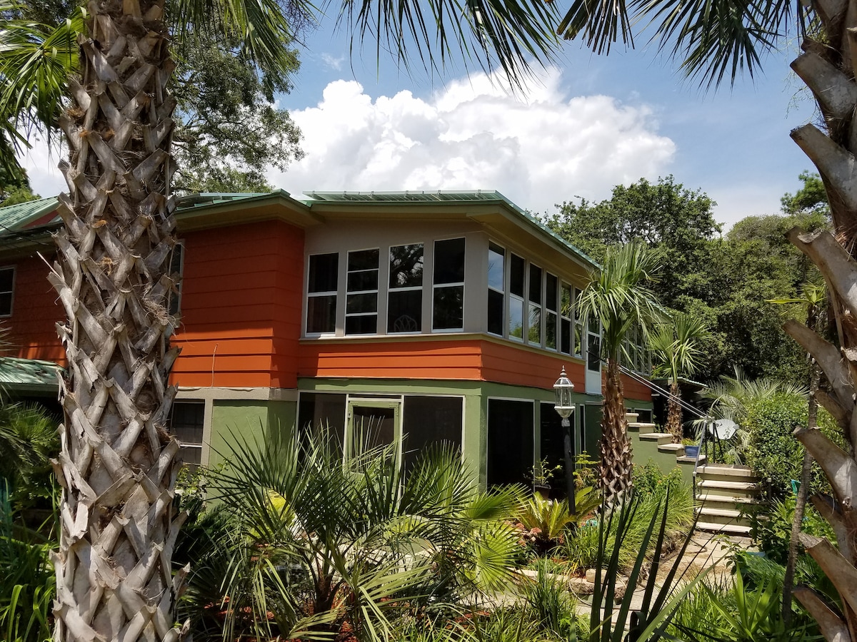 A vibrant two-story home is surrounded by lush greenery and palm trees. Large windows allow natural light to fill the interior, while a stairway leads to the entrance, enhancing the inviting outdoor space.