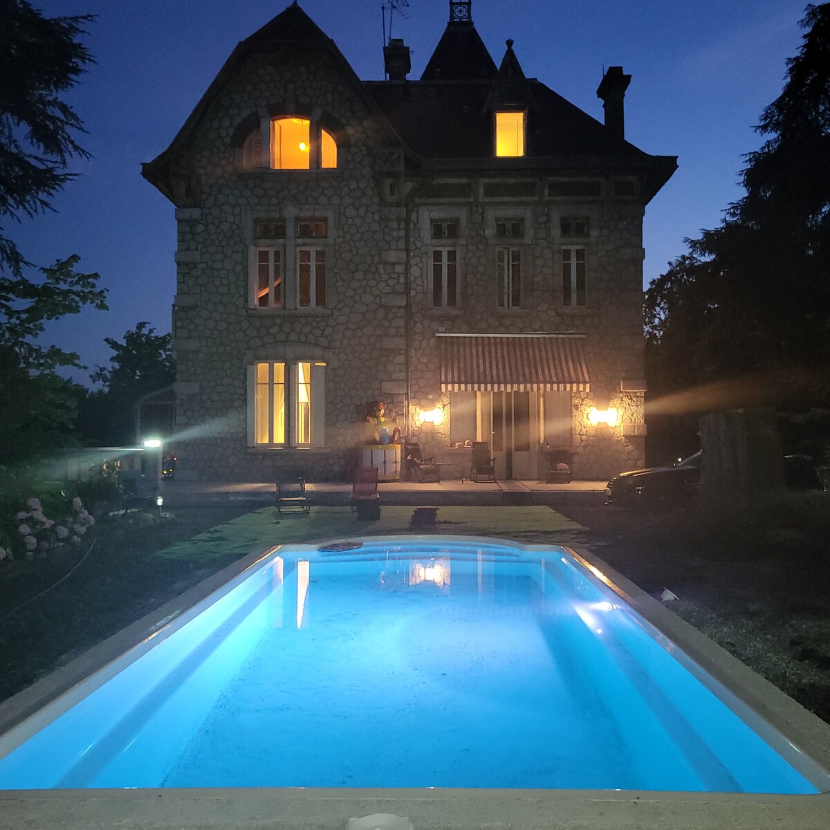 The garden of a grand stone mansion is showcased at night, with warm lights illuminating the windows. A well-maintained swimming pool in the foreground reflects the ambient glow, offering a serene atmosphere set against a backdrop of lush greenery.