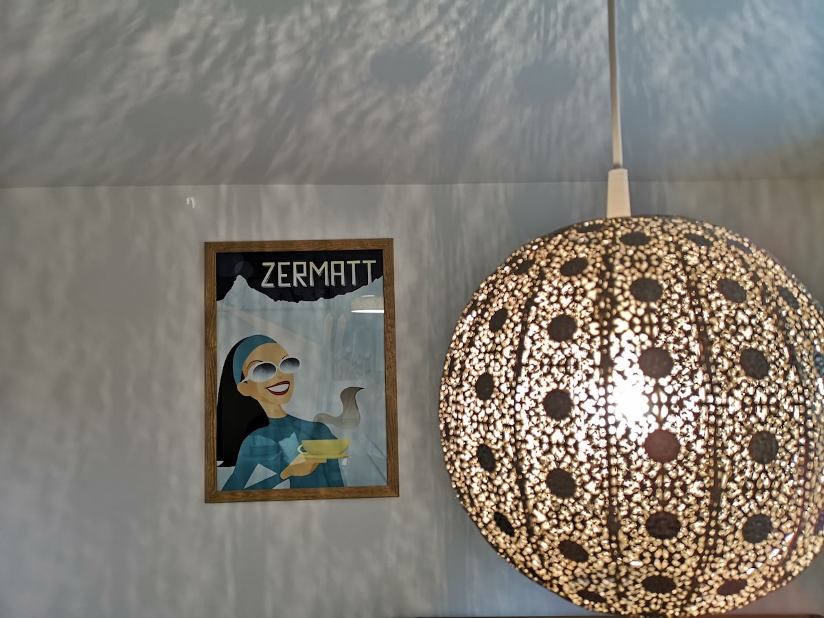 A decorative wall lamp with an intricate design hangs from the ceiling, casting soft patterns onto the wall. A framed artwork titled 'Zermatt' features a stylized image, adding character to the space. The colors are harmoniously integrated, contributing to the overall ambiance.