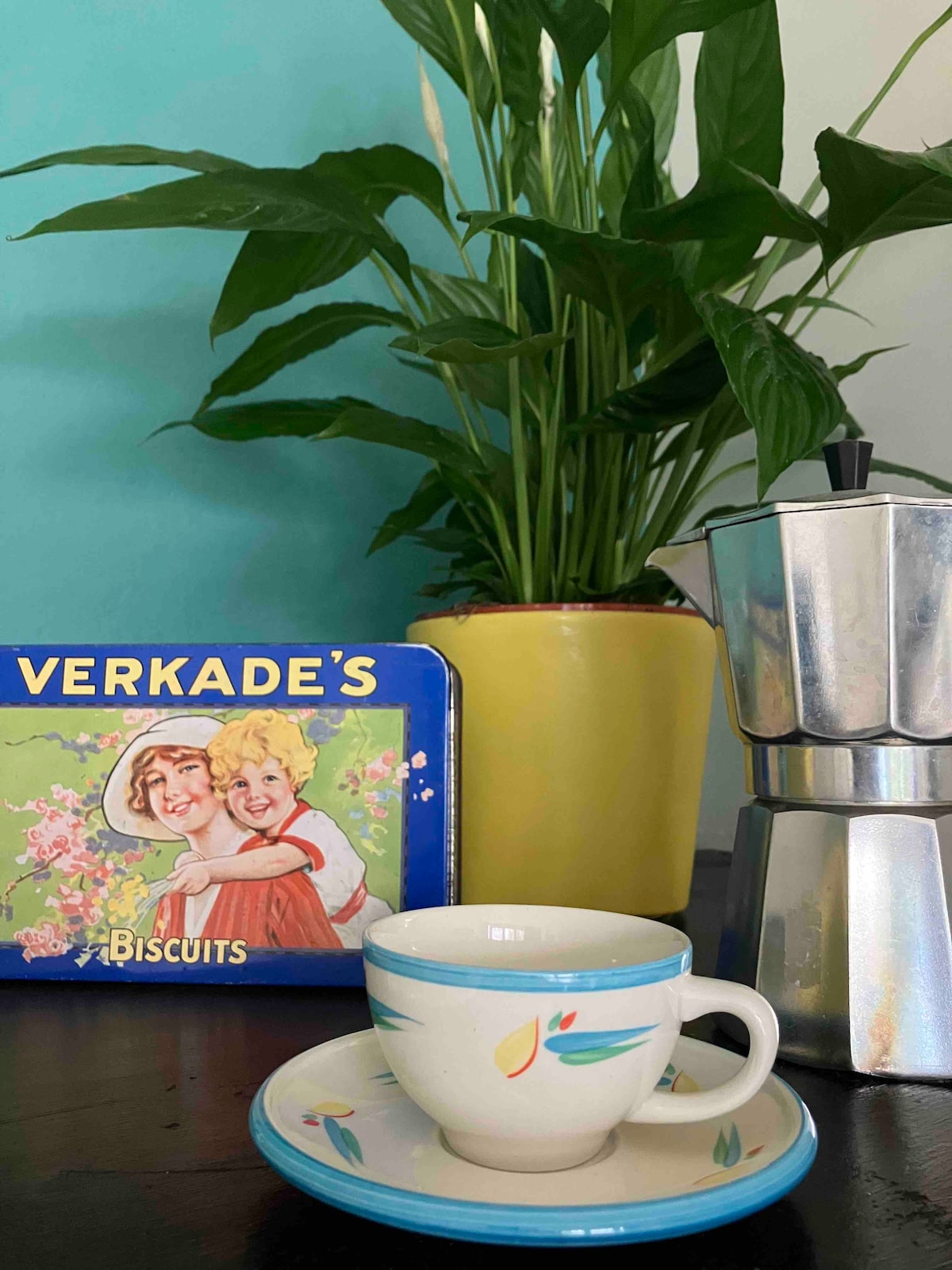 A vintage-style cup and saucer are positioned on a dark wooden surface, accompanied by a stainless steel coffee maker. A colorful tin of biscuits is displayed in the background, while a potted green plant adds a refreshing touch to the setting.