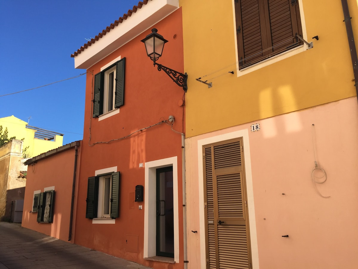 A charming building facade is presented, featuring vibrant colors of orange, yellow, and pink. Shuttered windows with dark frames add character, while a classic lantern hangs beside the entrance. The setting is in a quiet street, contributing to a serene atmosphere.