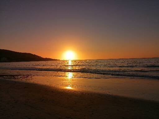 A sunset over the ocean is captured with warm hues of orange and yellow reflecting on the water's surface. The shoreline is visible, along with gentle waves lapping against the beach. A silhouette of a distant hill is observed in the background.