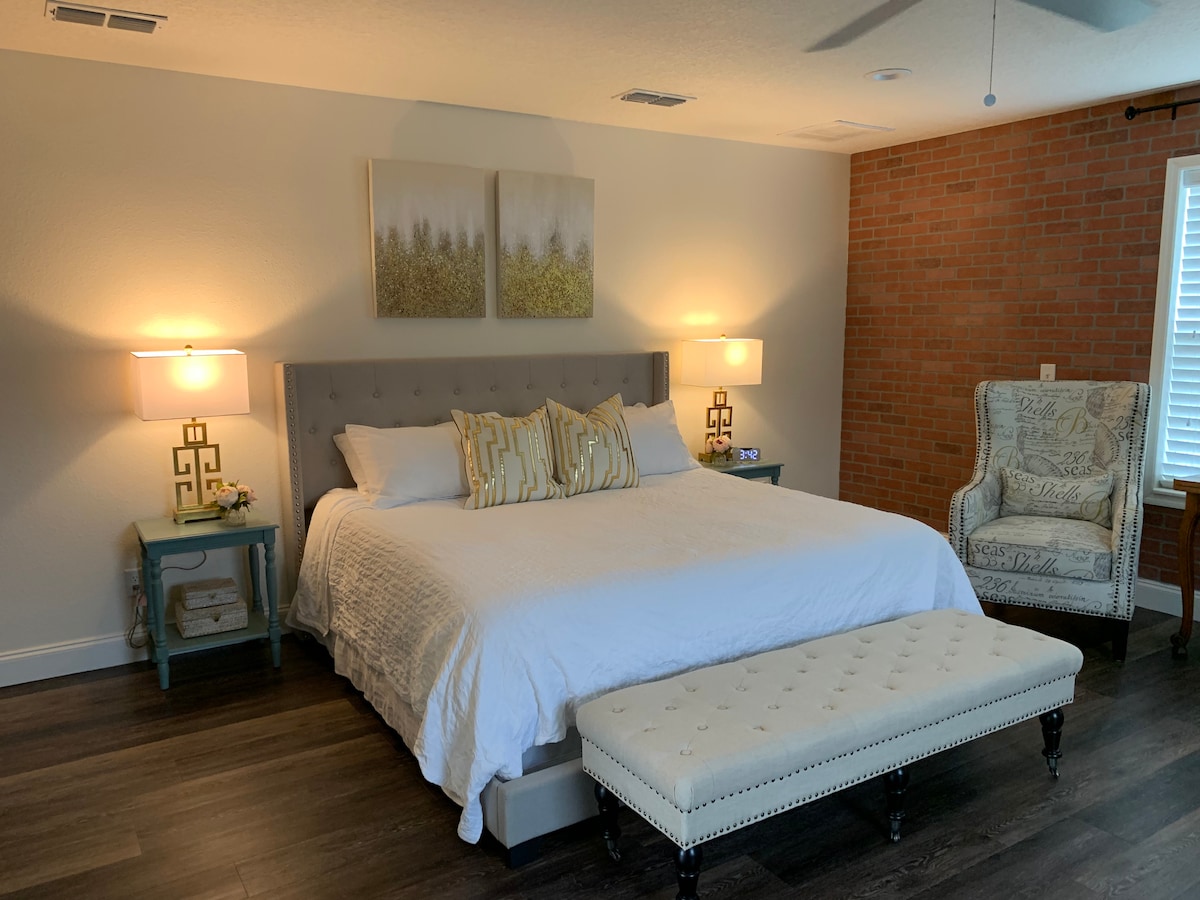 A spacious bedroom features a large bed with white bedding and decorative pillows. Two soft bedside lamps illuminate the area, complemented by a cozy armchair and a decorative bench at the foot of the bed. Exposed brick wall accents the room.
