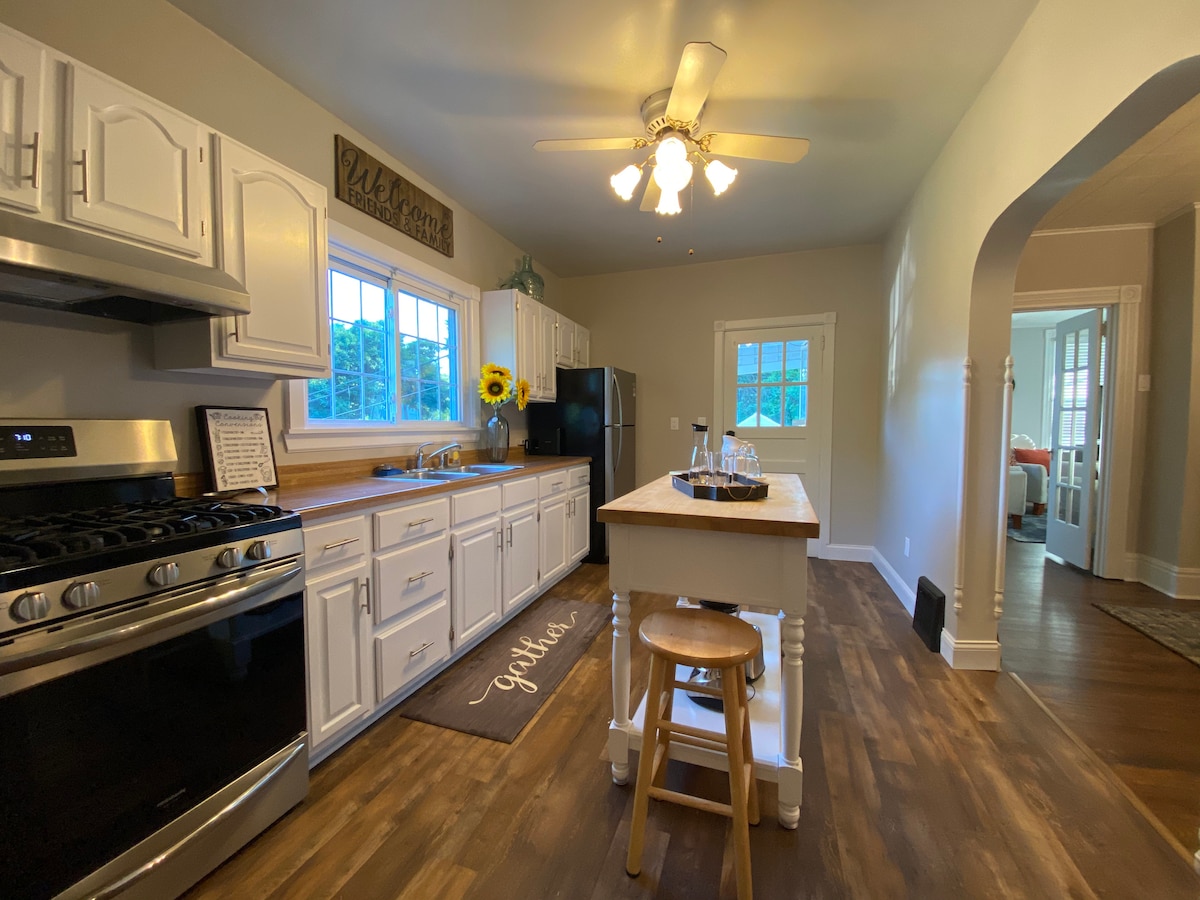 A well-appointed kitchen is highlighted with white cabinetry and stainless steel appliances. A central island offers additional workspace, complemented by a wooden countertop. Sunlight enters through multiple windows, illuminating a warm and inviting atmosphere. A cozy dining area is visible beyond an arched entryway.